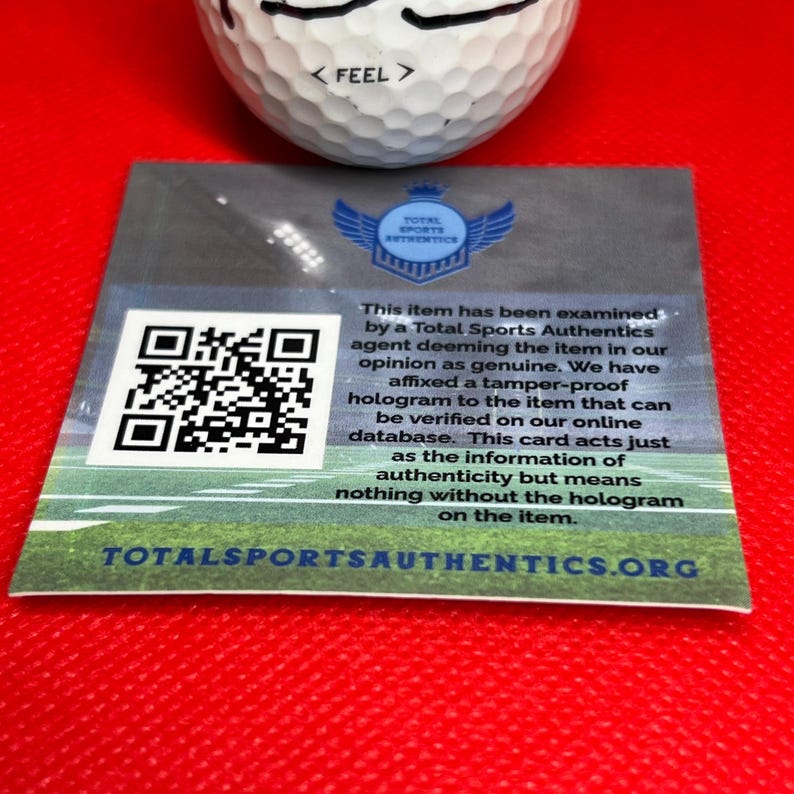 May include: A white golf ball with the word "< FEEL >" printed on it, resting above a rectangular card. The card features a QR code, a logo, and text that reads "TOTAL SPORTS AUTHENTICS". The card is on a red surface.