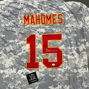 Peut inclure: Un maillot de camouflage gris et blanc avec le nom "MAHOMES" en lettres jaunes et rouges dans le haut du dos. Le numéro "15" est affiché en rouge avec un contour jaune au dos. Une signature est visible.