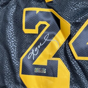 May include: A black athletic jersey with a textured, snakeskin-like pattern featuring a large yellow number 24 with black trim. A silver signature is written across the number, and a small rectangular holographic authentication sticker is attached.