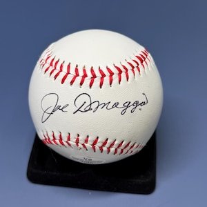 Op de afbeelding: Een witte lederen honkbal met rode stiksels en de handtekening van Joe DiMaggio in zwarte inkt in het midden. De bal staat op een zwarte vierkante displaystandaard en toont de authentieke handtekening en het textuur van het leer.