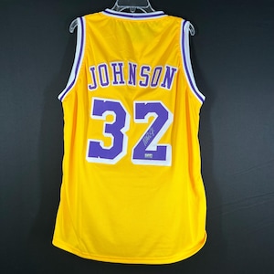 May include: A yellow basketball jersey with purple lettering and trim. The back of the jersey has the name "JOHNSON" and the number "32" on it. The jersey is signed in purple ink.