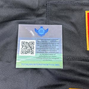May include: A black garment with a Total Sports Authentics authenticity card attached. The card features a QR code, a logo with wings, and text stating the item has been examined and is deemed genuine.