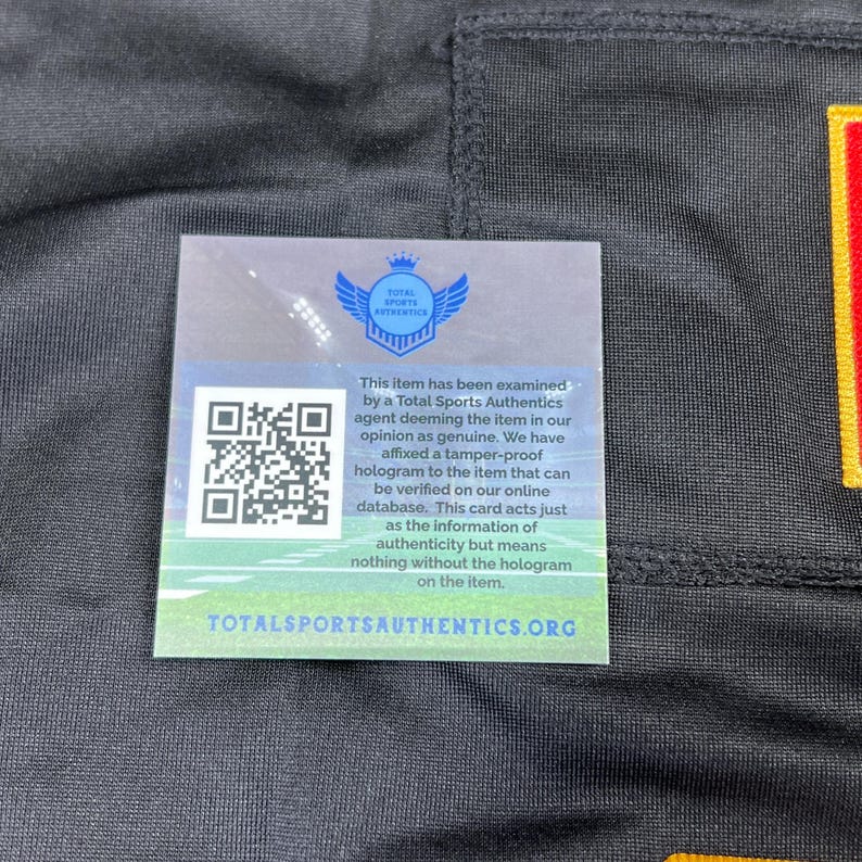 May include: A black garment with a Total Sports Authentics authenticity card attached. The card features a QR code, a logo with wings, and text stating the item has been examined and is deemed genuine.