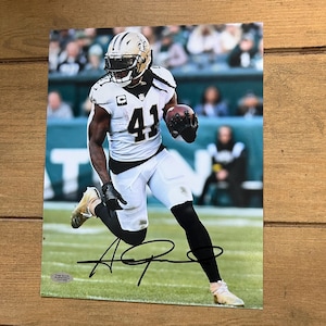 May include: A signed photograph of a football player in a white and black uniform, number 41, running on a green field. The player is wearing a gold helmet and holding a football. The background shows a stadium with spectators.