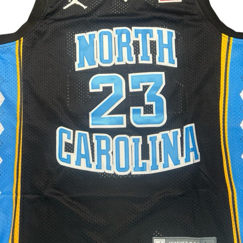 May include: Black basketball jersey with the words "NORTH 23 CAROLINA" in light blue with white trim. The jersey has a mesh design and a yellow stripe down the side with a light blue panel. The Jordan logo is visible.
