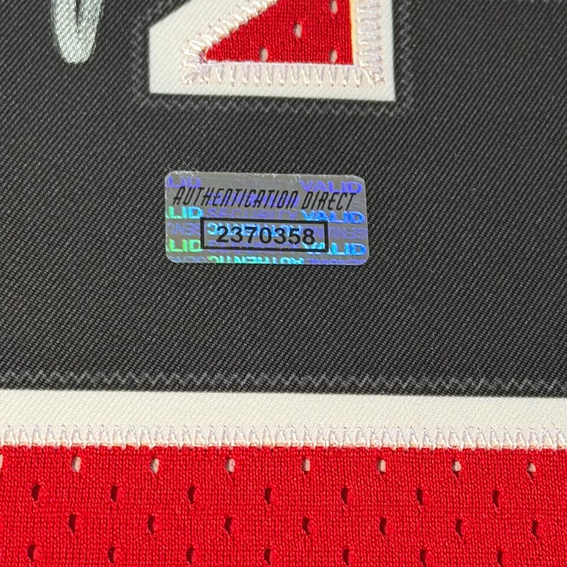 Puede incluir: Una camiseta deportiva negra y roja con un parche de n&uacute;mero cosido en blanco y una pegatina de autenticaci&oacute;n hologr&aacute;fica rectangular con el texto AUTHENTICATION DIRECT y el n&uacute;mero 2370358 en la secci&oacute;n de tela negra.