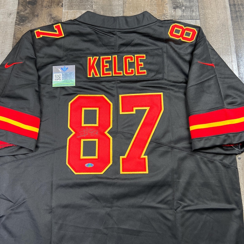 May include: Black jersey with red and yellow accents. The jersey features the name "KELCE" in red lettering and the number "87" in red with a yellow outline. The number "7" is visible on the shoulders. A Nike logo is present.