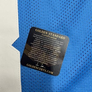 K&ouml;nnte beinhalten: Nahaufnahme eines blauen Mesh-Stoffs mit wei&szlig;em Besatz. Ein schwarzes und goldenes Zertifizierungsetikett ist angebracht, mit dem Text "GOLDEN STANDARD Certification" und "GOLDENCERTS.COM".