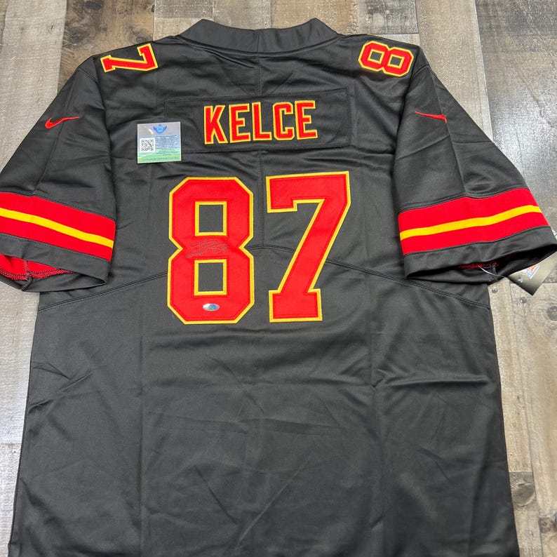 May include: A black football jersey with red and yellow accents. The jersey features the name "KELCE" and the number "87" in red with a yellow outline. The sleeves have red and yellow stripes.