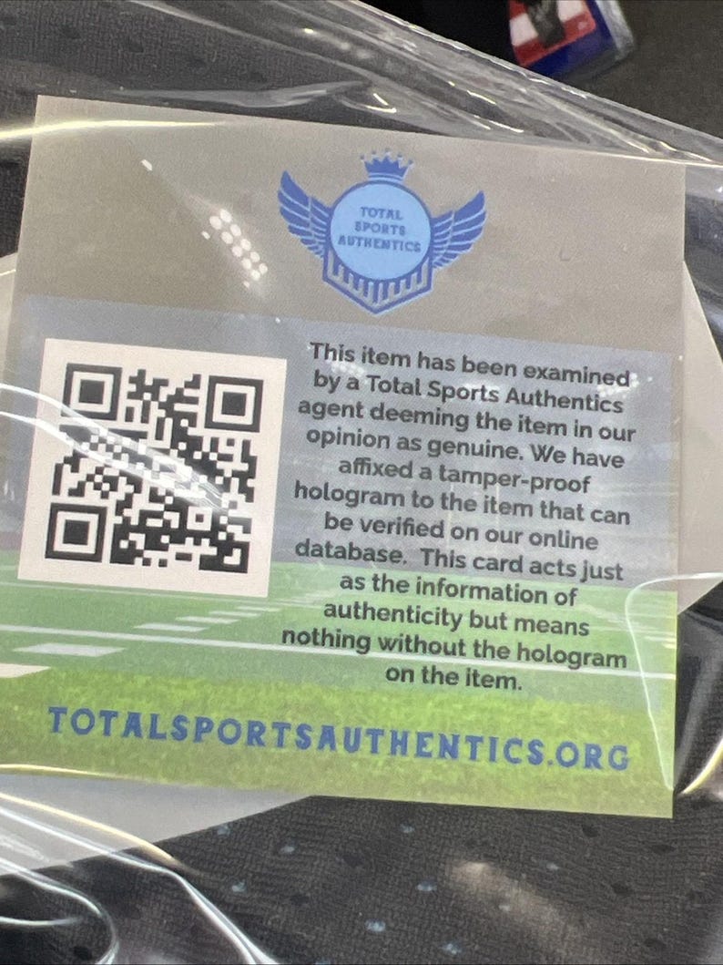 May include: A clear plastic bag containing a white and green Total Sports Authentics authenticity card. The card has a QR code, a blue logo with wings, and text stating the item has been examined and is deemed genuine. The website TOTALSPORTSAUTHENTICS.ORG is also visible.
