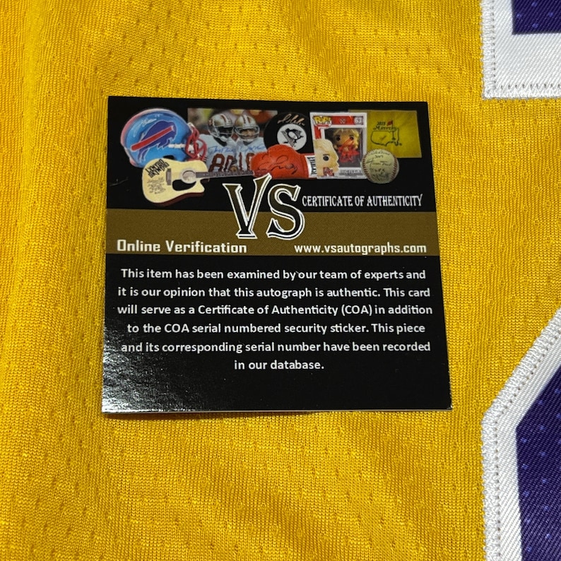 May include: A yellow jersey with a black and white certificate of authenticity. The certificate includes the text "VS" and "Certificate of Authenticity". The certificate also has the text "Online Verification" and "www.vsautographs.com".