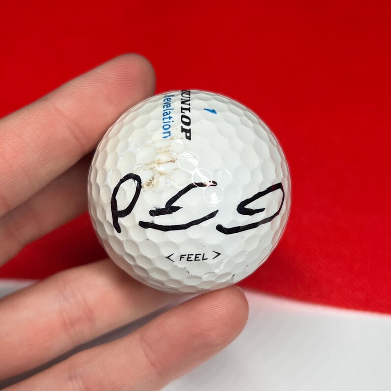 May include: A white Dunlop golf ball with black markings and the word "FEEL" printed below the markings. The golf ball has a dimpled surface and the number 1 in blue.