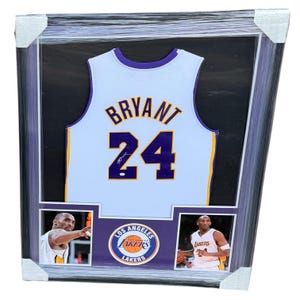 May include: A framed white Los Angeles Lakers basketball jersey with the name BRYANT and number 24 in purple and gold, featuring a signature on the number. Below are two photographs of a player and a circular Los Angeles Lakers team logo.