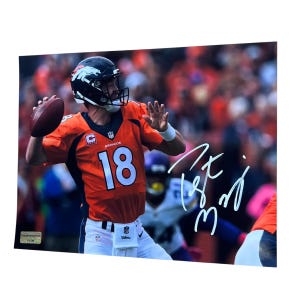May include: A framed photograph of a football player in an orange and blue uniform throwing a football. The player is wearing a black helmet with a white horse logo. The jersey number 18 is visible. The photograph is signed in white ink.