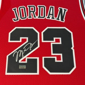 Puede incluir: Una camiseta de baloncesto de malla roja con el nombre JORDAN en letras negras con borde blanco sobre el n&uacute;mero 23 en negro con borde blanco. Una firma plateada cruza el n&uacute;mero 23 y se ve una peque&ntilde;a etiqueta de autenticaci&oacute;n.
