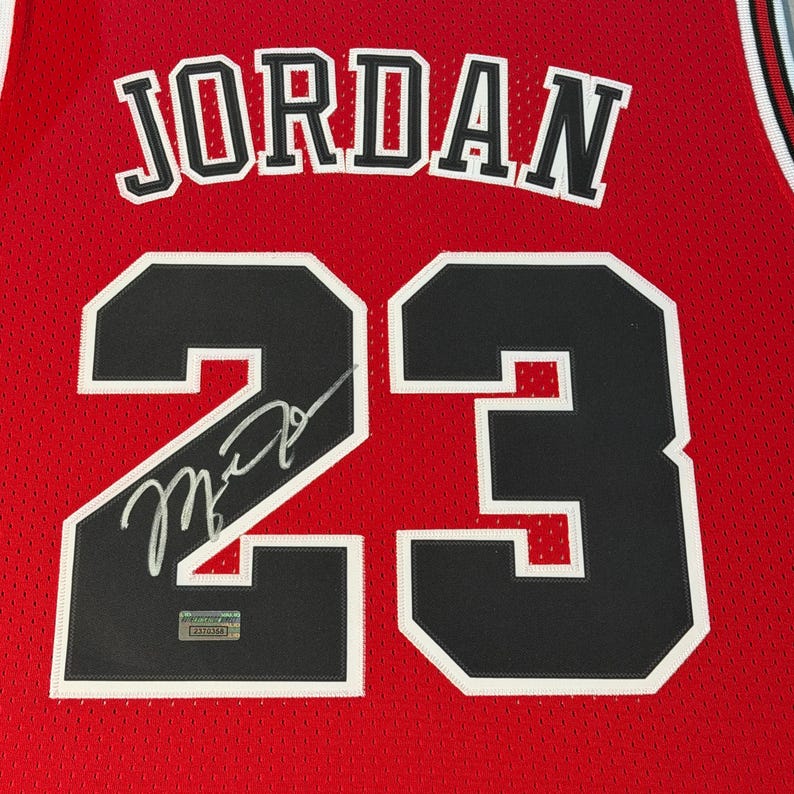 Puede incluir: Una camiseta de baloncesto de malla roja con el nombre JORDAN en letras negras con borde blanco sobre el n&uacute;mero 23 en negro con borde blanco. Una firma plateada cruza el n&uacute;mero 23 y se ve una peque&ntilde;a etiqueta de autenticaci&oacute;n.