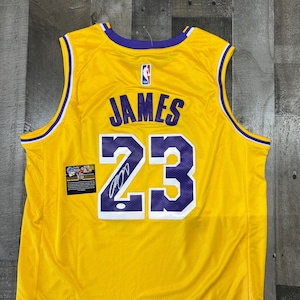 May include: A yellow basketball jersey with purple and white accents. The jersey features the name "JAMES" and the number "23" in purple. An NBA logo is visible, along with a small patch with text and a signature.