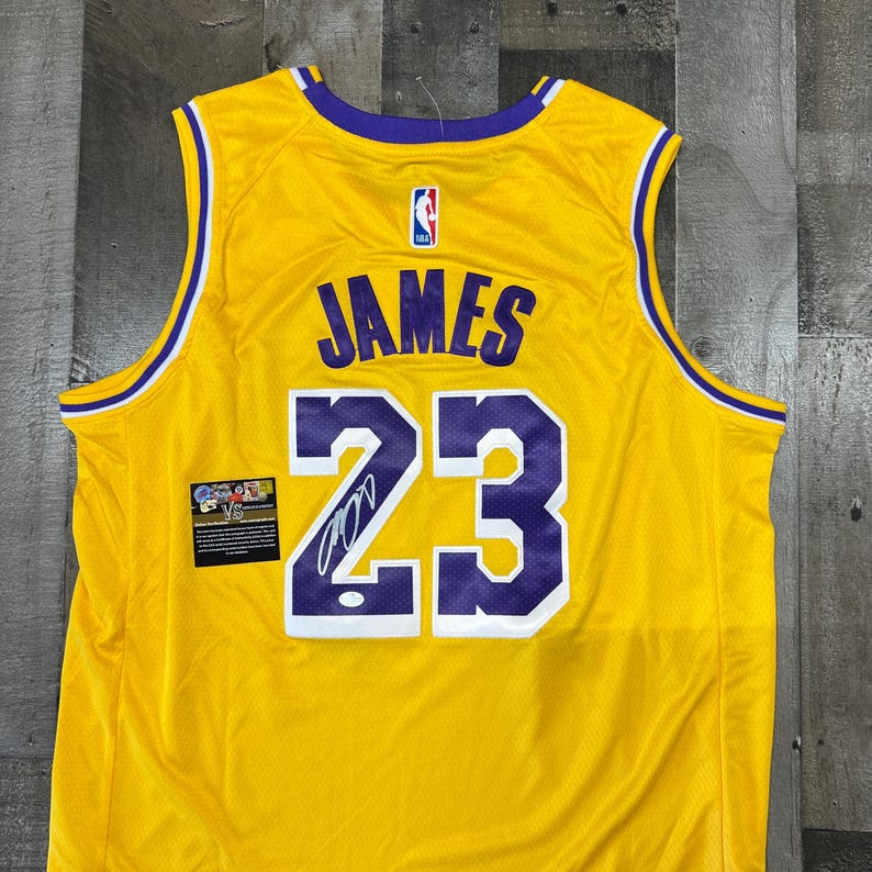 May include: A yellow basketball jersey with purple and white accents. The jersey features the name "JAMES" and the number "23" in purple. An NBA logo is visible, along with a small patch with text and a signature.
