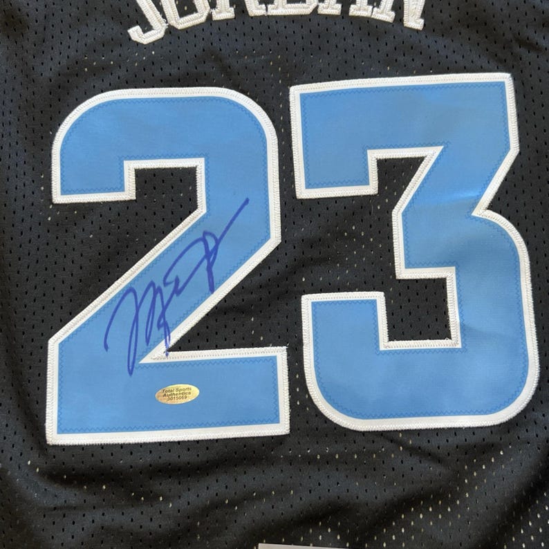 May include: Black mesh jersey with the number 23 in light blue and white. The jersey has a signature in blue ink on the number 23. A small, round sticker is visible near the signature. The jersey has the name "JORDAN" in white letters.