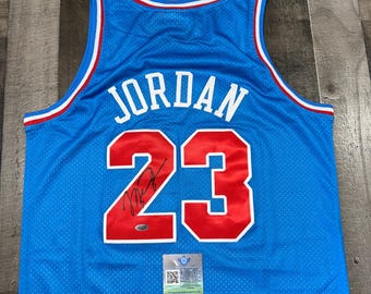 May include: A blue basketball jersey with the name "JORDAN" in white letters and the number "23" in red. The jersey has a red and white trim and a signature on the number. A small authentication sticker is visible.