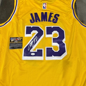 May include: A yellow basketball jersey with purple accents, featuring the name "JAMES" and the number "23" in purple. The jersey has a signature and a small authentication sticker. The NBA logo is visible.