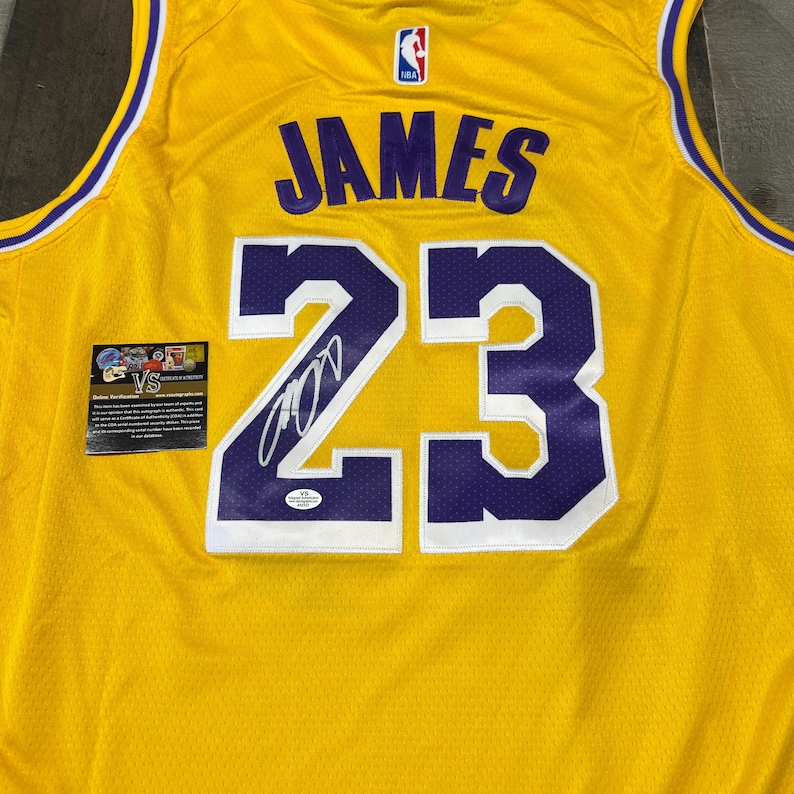 May include: A yellow basketball jersey with purple accents, featuring the name "JAMES" and the number "23" in purple. The jersey has a signature and a small authentication sticker. The NBA logo is visible.