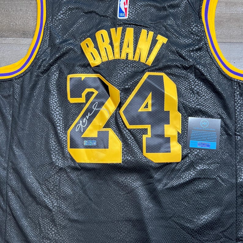 May include: A black basketball jersey with a snakeskin-like texture, featuring the name BRYANT and the number 24 in yellow with black outlines. The jersey includes a signature on the number 24, an NBA logo at the neckline, and yellow and purple trim.