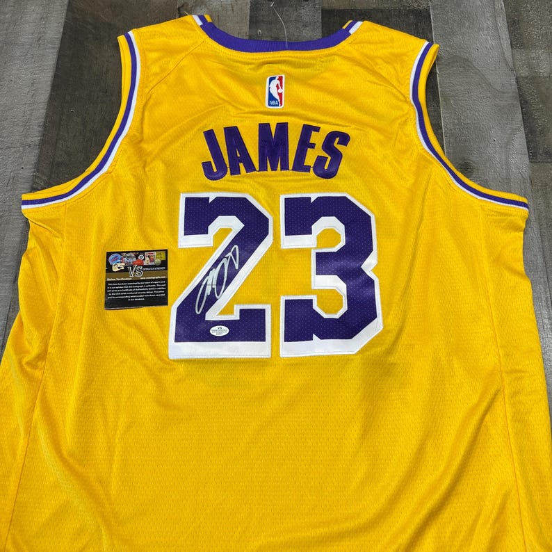 May include: A yellow basketball jersey with purple and white accents. The jersey features the name "JAMES" and the number "23" in purple, along with an autograph. A small patch is visible on the left side.