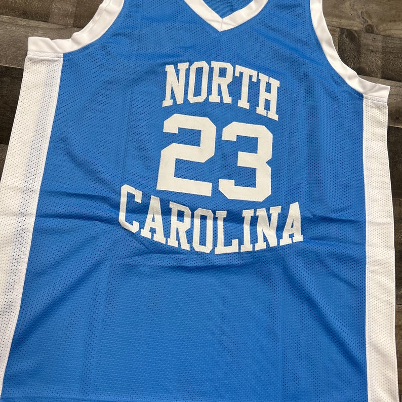 K&ouml;nnte beinhalten: Hellblaues Basketballtrikot mit den wei&szlig;en Worten "NORTH 23 CAROLINA". Das Trikot hat einen V-Ausschnitt und wei&szlig;e Verzierungen. Der Stoff hat eine Mesh-Struktur. Es ist ein Sportbekleidungsst&uuml;ck.
