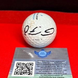 May include: A white golf ball with black markings and the word "FEEL" printed on it. The ball is signed with black ink. A card with a QR code and text from Total Sports Authentics is in the foreground.