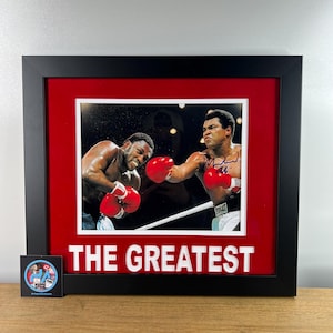 Può includere: Stampa incorniciata con un incontro di boxe con due pugili con guantoni rossi. La stampa è ambientata su un passe-partout rosso e incorniciata in nero. Il testo "THE GREATEST" è visualizzato in lettere bianche.