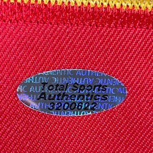 May include: Close-up of a red fabric with a yellow border. A holographic oval sticker is affixed to the fabric, displaying the text "Total Sports Authentics" and a serial number. The fabric has a textured appearance.