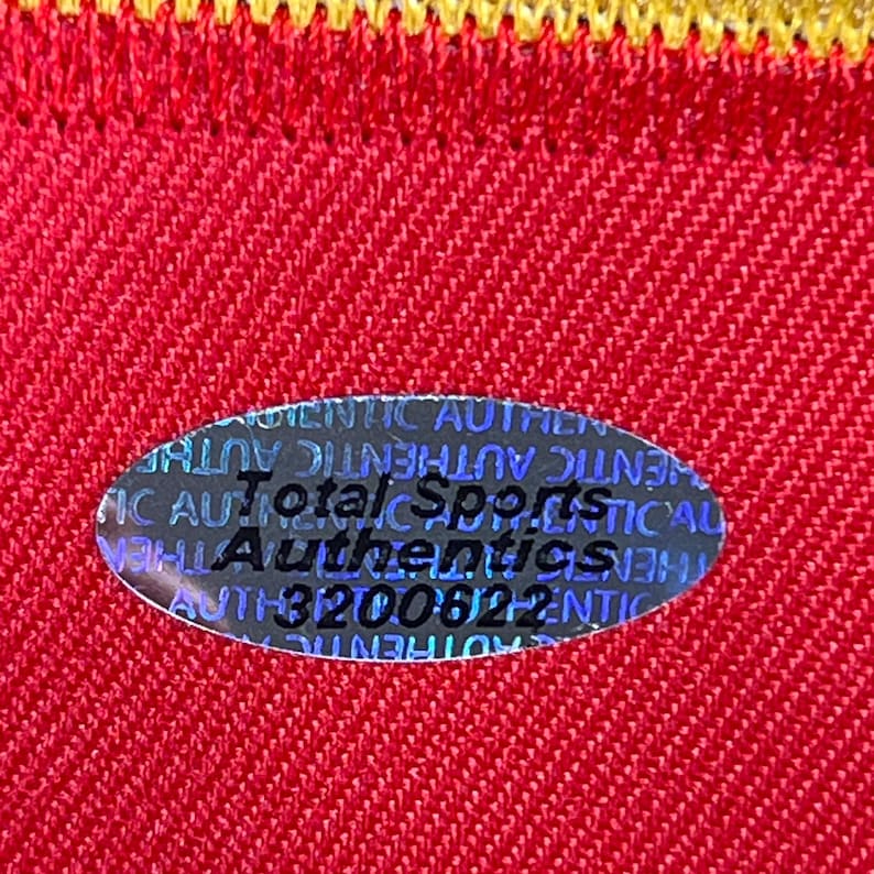 May include: Close-up of a red fabric with a yellow border. A holographic oval sticker is affixed to the fabric, displaying the text "Total Sports Authentics" and a serial number. The fabric has a textured appearance.