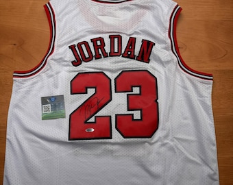 Puede incluir: Una camiseta de baloncesto blanca con ribetes rojos y negros en el cuello y las sisas. La espalda muestra el nombre JORDAN y el número 23 en rojo con bordes negros. Una firma negra aparece sobre el número 23, junto a una etiqueta de autenticidad.