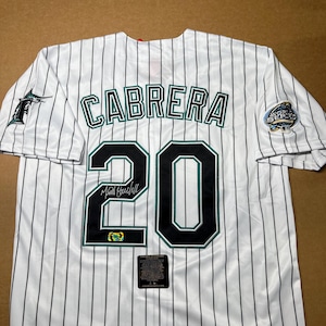 May include: A white baseball jersey with black pinstripes, featuring the name CABRERA and the number 20 in black with teal outlines on the back. The jersey includes a Marlins logo on the right sleeve, a 2003 World Series patch on the left, and a signature.
