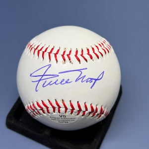 Pode incluir: Uma bola de beisebol branca com costuras vermelhas apresentando um autógrafo em tinta azul no centro. Um pequeno adesivo oval na parte inferior exibe o texto VS Autograph Authentication, o site vsautographs.com e o número A50784.
