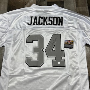 Pode incluir: Camisola de futebol branca com o nome "JACKSON" nos ombros e o número "34" em cinza. A camisola tem uma assinatura e um pequeno autocolante. A camisola é feita de um material de malha.
