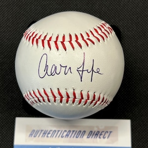 May include: A white baseball with red stitching and a handwritten signature in blue ink. The baseball is on a black surface, with a white and blue label at the bottom that reads "AUTHENTICATION DIRECT."