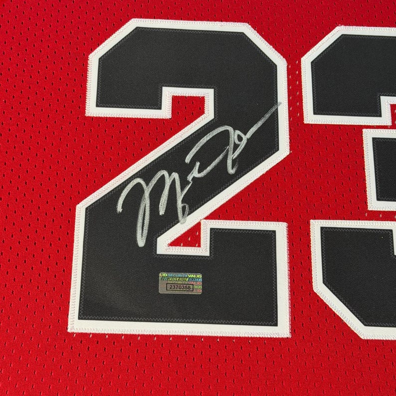 Puede incluir: Una camiseta deportiva de malla roja con el n&uacute;mero 23 en negro y borde blanco. Un aut&oacute;grafo plateado est&aacute; escrito sobre el n&uacute;mero 23. Una peque&ntilde;a pegatina de autenticaci&oacute;n rectangular con el texto LID SECURITY VALID est&aacute; adherida al n&uacute;mero.