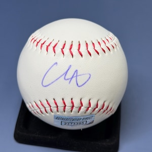 May include: A white leather baseball featuring red stitching and a blue ink autograph on the surface. A rectangular authentication sticker is affixed to the lower portion of the ball, displaying the text AUTHENTICATION DIRECT and the number 2373395.