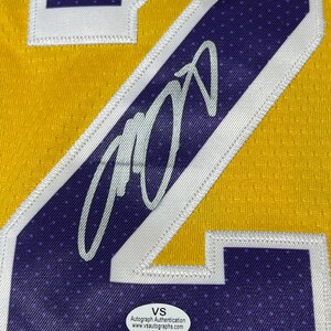 May include: Close-up of a yellow and purple basketball jersey with a large number 24. The jersey features a silver autograph and a white and purple patch. A VS Autograph Authentication sticker is visible.