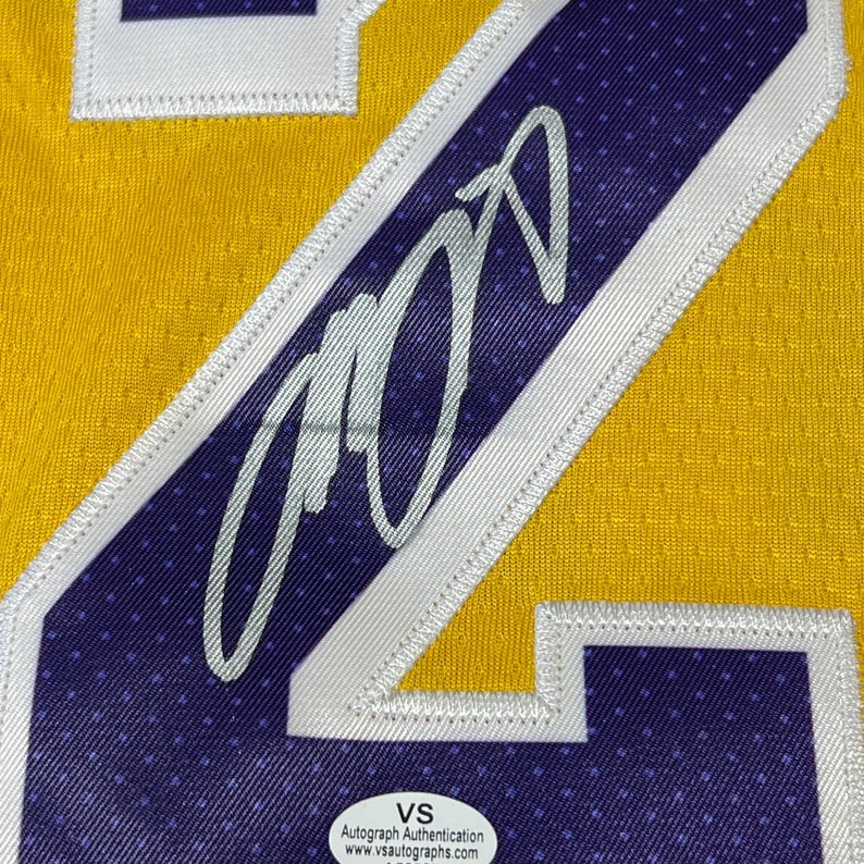 May include: Close-up of a yellow and purple basketball jersey with a large number 24. The jersey features a silver autograph and a white and purple patch. A VS Autograph Authentication sticker is visible.