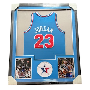 Pode incluir: Uma camisola de basquetebol azul-clara emoldurada com o nome JORDAN e o número 23 a vermelho com contorno branco. A camisola apresenta uma assinatura preta e um pequeno autocolante de autenticidade. Abaixo, duas fotos de ação e um logótipo.