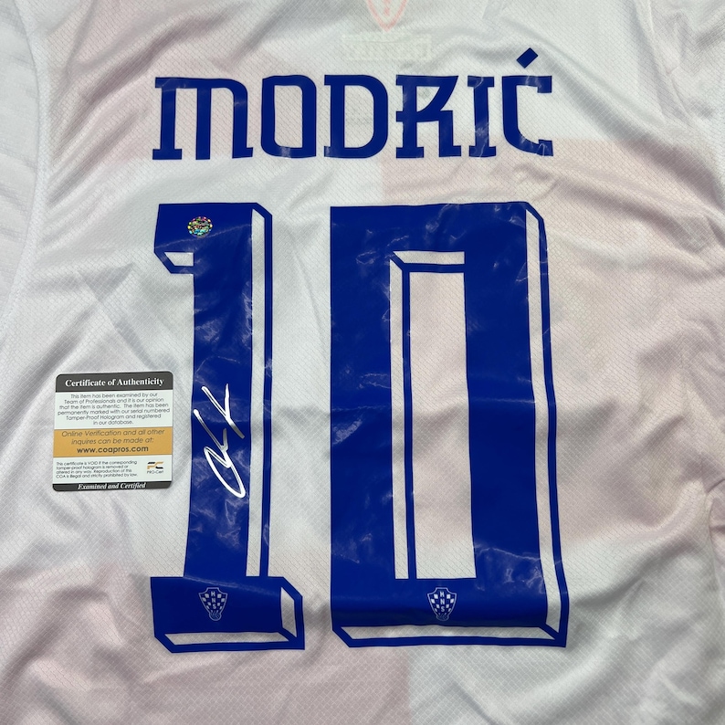 Puede incluir: Camiseta de f&uacute;tbol blanca con el nombre "MODRIĆ" en letras azules en bloque sobre el n&uacute;mero "10". El n&uacute;mero tambi&eacute;n es azul. Se ve un certificado de autenticidad. La camiseta tiene una firma en el n&uacute;mero.