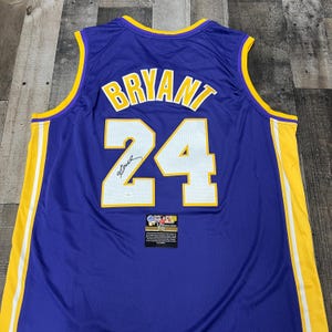 May include: A purple basketball jersey featuring the name BRYANT and the number 24 in white with yellow trim. The jersey includes a black signature on the number 24 and a small rectangular authentication label with text at the bottom centre.