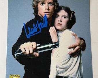 May include: A glossy photograph featuring Luke Skywalker in a black shirt holding a silver lightsaber and Princess Leia in a white dress, with a blue ink autograph across the center and a gold holographic sticker in the bottom left corner.