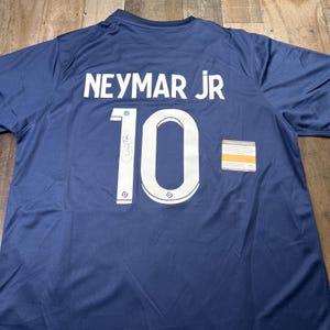 Pode incluir: Camisola de futebol azul marinho com o nome "NEYMAR JR" e o número "10" em branco. A camisola tem uma assinatura e um pequeno certificado. O tecido é leve e respirável.