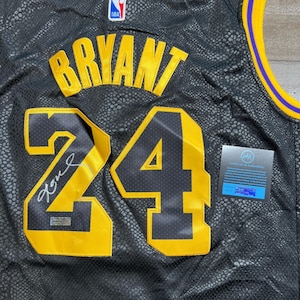 May include: A black basketball jersey with a textured snakeskin pattern, featuring the name BRYANT and the number 24 in yellow and black. The jersey includes an NBA logo, a silver autograph on the number 2, and a certificate of authenticity card.