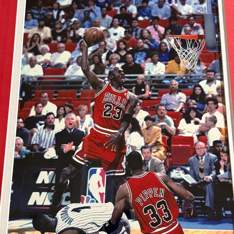 Puede incluir: Una fotograf&iacute;a deportiva que muestra a Michael Jordan con una camiseta roja de los Chicago Bulls con el n&uacute;mero 23, saltando para encestar. Scottie Pippen con una camiseta roja con el n&uacute;mero 33 est&aacute; cerca, mientras un jugador con camiseta a rayas est&aacute; en el suelo.