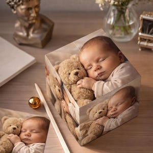 Op de afbeelding: Een fotoblok met een slapende baby die een teddybeer knuffelt. De afbeelding wordt op meerdere zijden van de kubus herhaald, waardoor een 3D-effect ontstaat. Een ingelijste afdruk van dezelfde afbeelding is ook zichtbaar.
